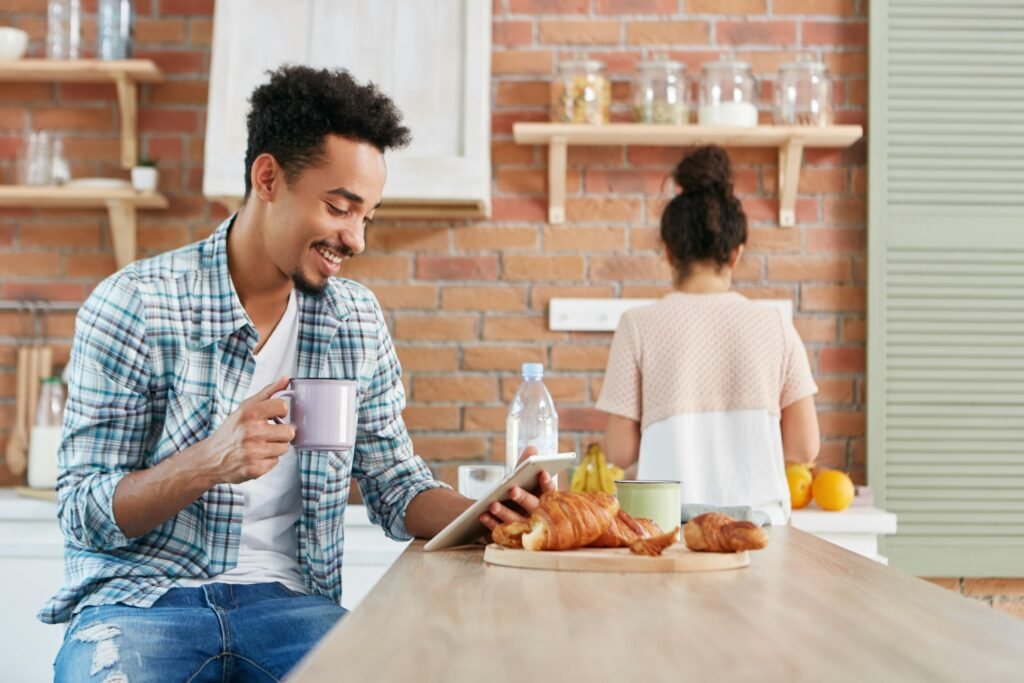 Cheerful mixed race male watches comedy on tablet, uses free internet connection, drinks coffee and